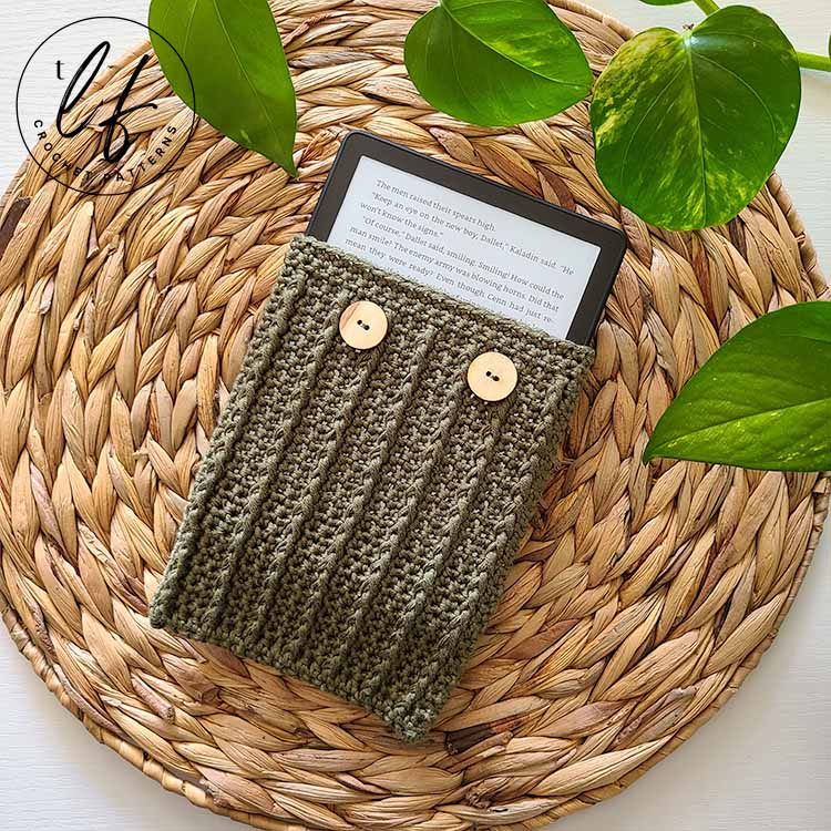 This image shows a finished crochet kindle cover placed three fourths of the way onto a kindle, so that both the kindle and the cover can be seen. The device and cover are laid flat on a jute placemat with a plant peaking in at the top.