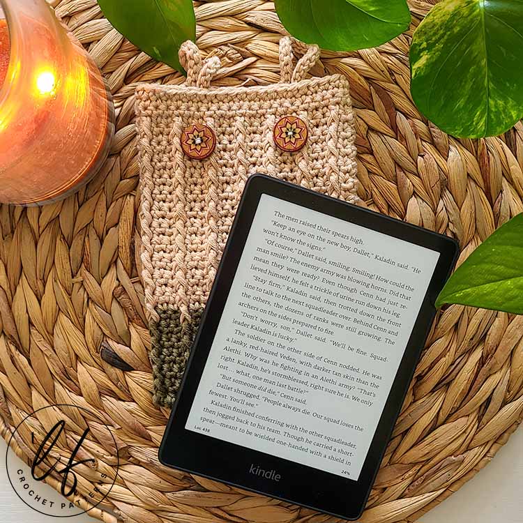 This image shows a finished crochet kindle cover with a kindle placed ion top and slightly diagonal to the cover. The device and cover are laid flat on a jute placemat with a plant peaking in at the top.