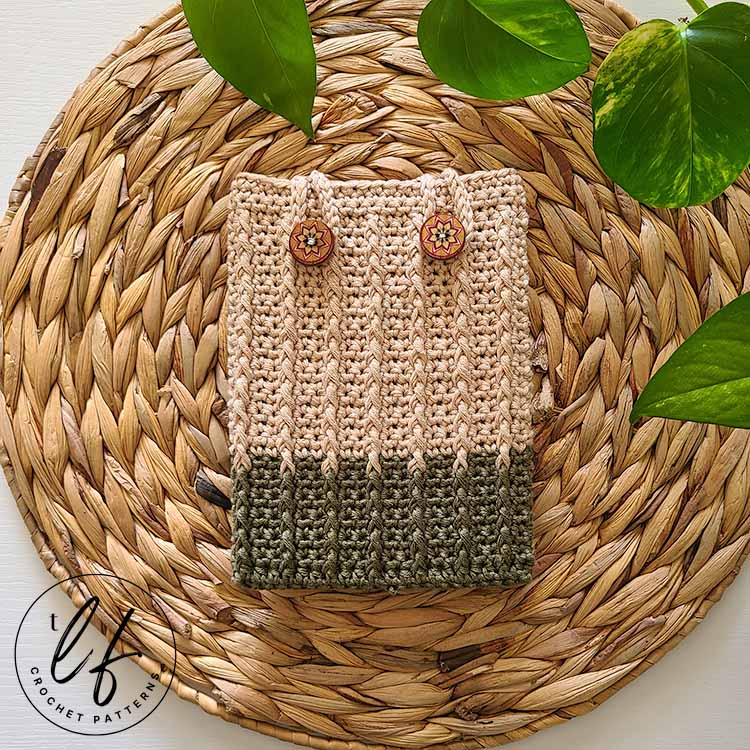 This image shows a finished crochet kindle sleeve with a kindle placed inside and the loops secured. The device and cover are laid flat on a jute placemat with a plant peaking in at the top.