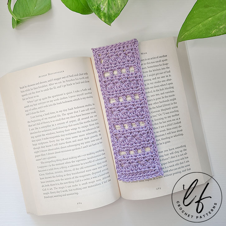 This image shows the finished project made from this Lace Crochet Bookmark Pattern in an open, paperback book that is medium sized. The bookmark is made in a lavender color.