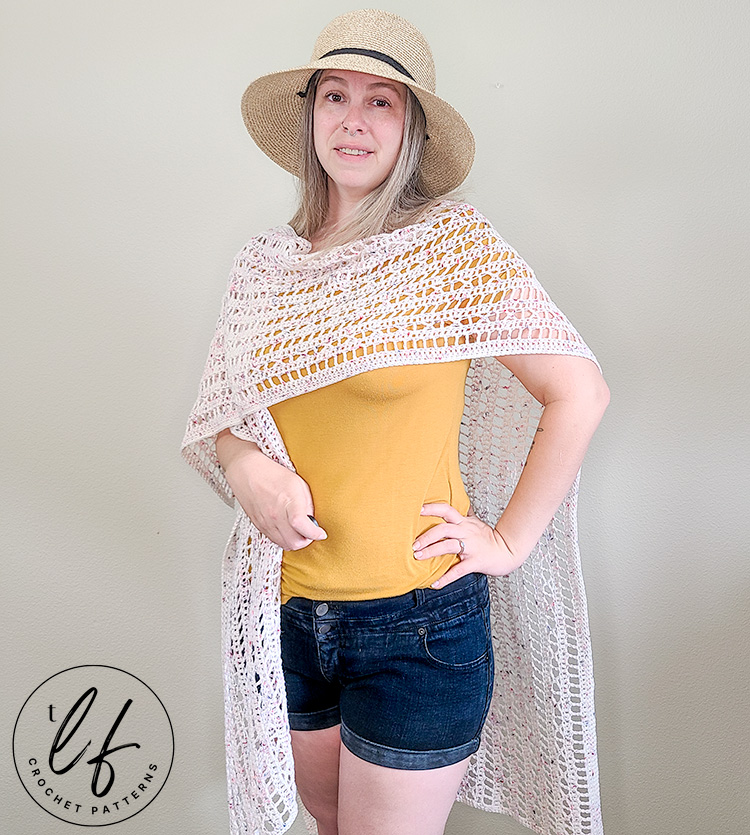 This image shows a woman wearing a finished ruana from this Crochet Ruana Pattern. The woman is standing in front of a blank wall and the picture is taken from the front and she has draped one of the front panels over her shoulder to show another way the ruana can be styled.