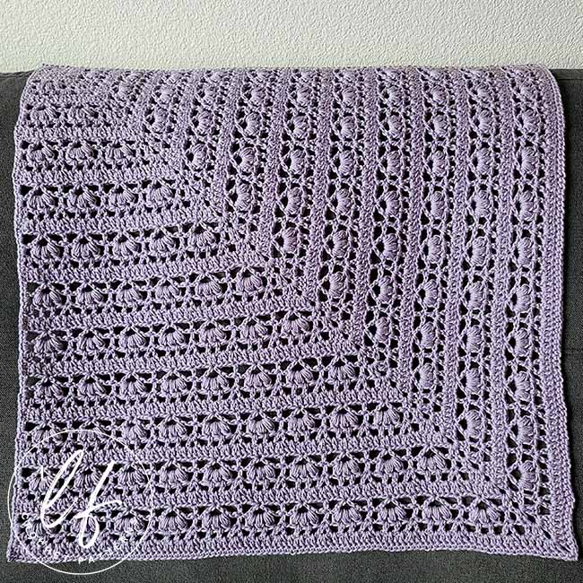 This image shows a finished sample of the flower square crochet pattern. The pattern has been worked in lavender yarn and is shown draped over a dark gray couch.