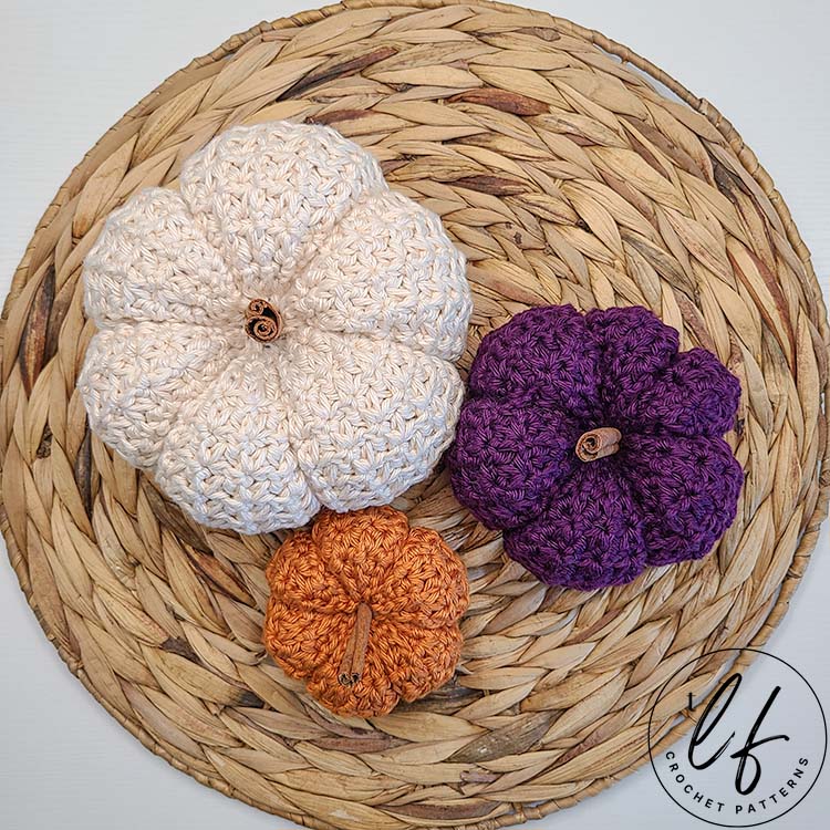 This image shows 3 pumpkins made from this crochet pumpkin pattern - one in each size. The large is made in cream, the small is made in purple and the tiny is made in orange. The pumpkins are shown sitting on a woven jute placemat. The picture is taken from the top down.