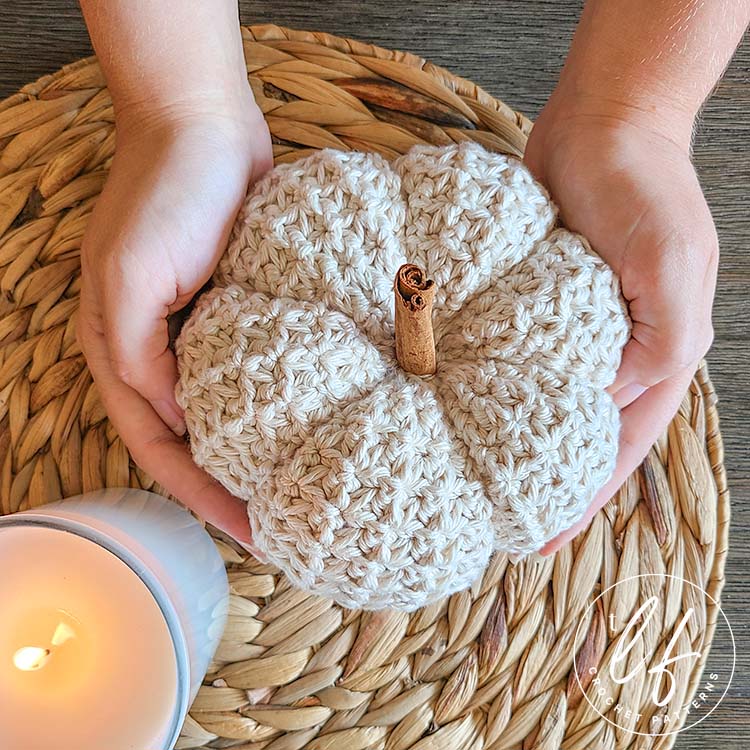 This image shows 1 pumpkin made from this crochet pumpkin pattern - the large size in cream.  The pumpkin is shown held by two hands a woven jute placemat with a white candle, as if in the process of being placed. The picture is taken from the top down.