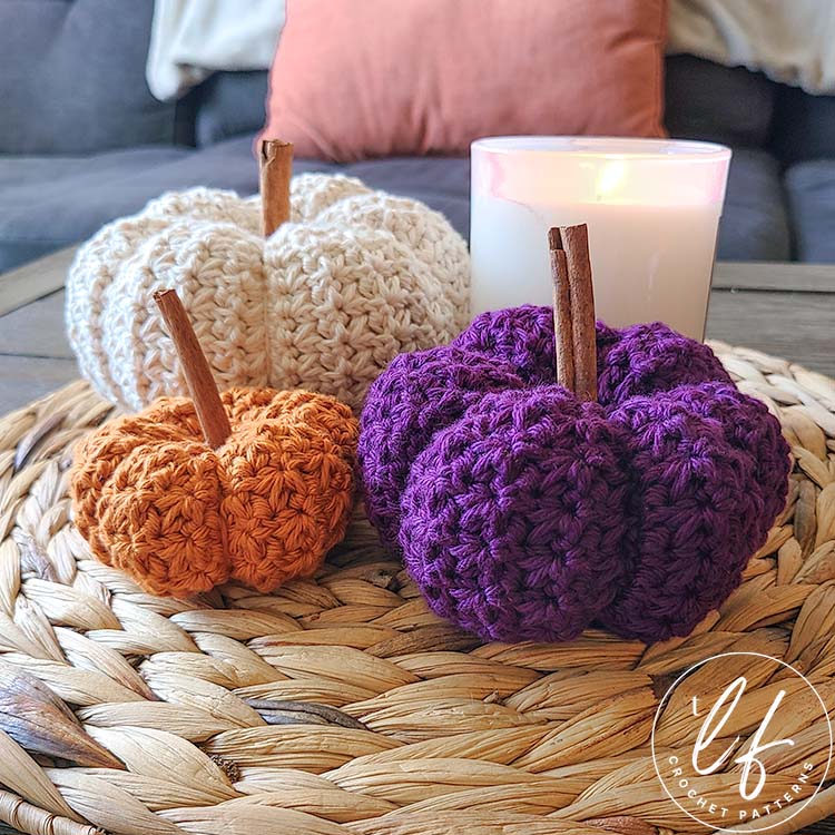 This image shows 3 pumpkins made from this crochet pumpkin pattern - one in each size. The large is made in cream, the small is made in purple and the tiny is made in orange. The pumpkins are shown sitting on a woven jute placemat with a white candle. The picture is taken from the side.