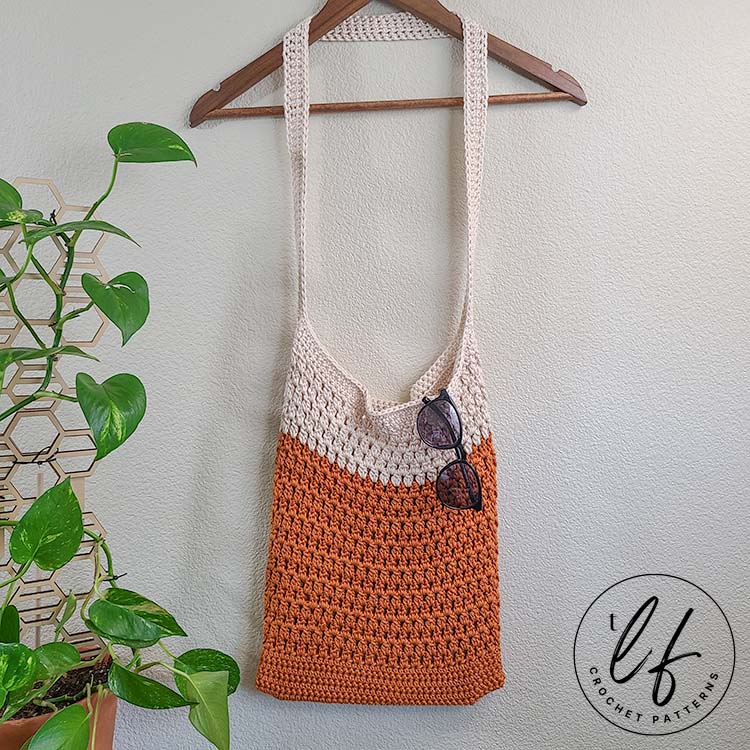 This image shows a finished bag from this crochet tote bag pattern. It is made in orange and cream and is hanging from a clothes hanger with sunglasses hanging from the top of the bag. 