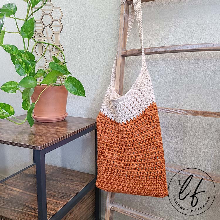 This image shows a finished bag from this crochet tote bag pattern. It is made in orange and cream and is hanging from a blanket ladder. There is also a side table, a plant and a candle.