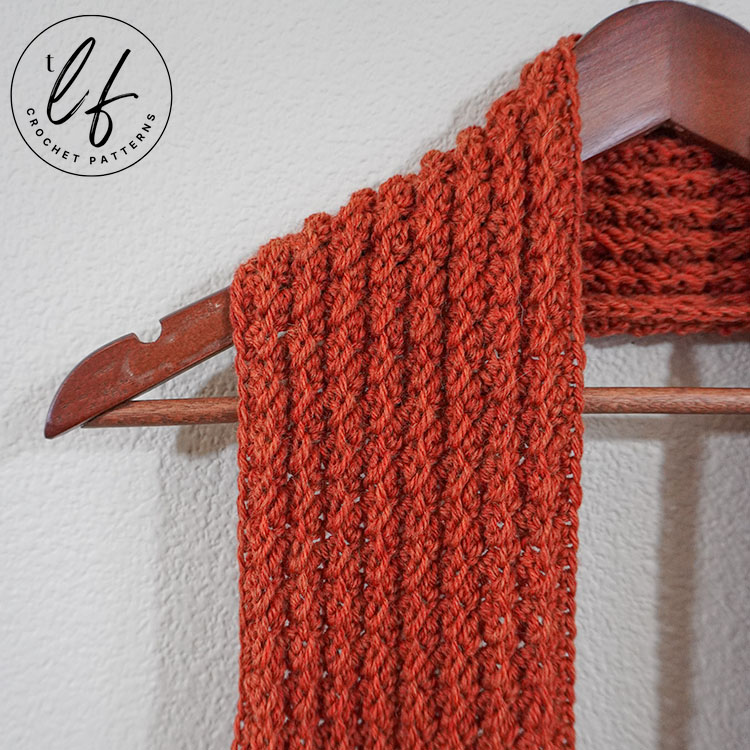 This image is a close up of the crochet scarf pattern sample. The scarf is hanging on a wooden clothes hanger on a white wall. The scarf is draped on the hanger as one would drape a scarf on their shoulders. The image is cropped and close up to show detail.